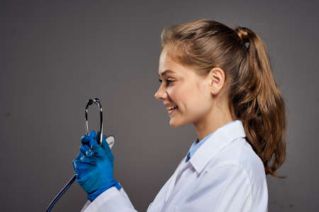 Woman holds a stethoscope in a medical dressing gown on a dark gray background, portrait, smile,...