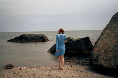 Young woman in a blue dress is standing on the beach near the rocks.の写真素材