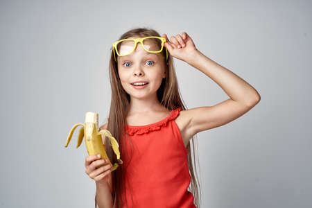 A little girl with long hair straightens glasses and holds a banana.の写真素材