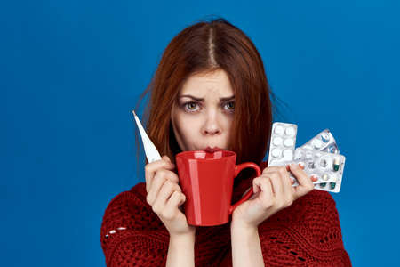 a girl with red hair, a mug and a sweater is holding a lot of pills and a thermometer on a blue background.の写真素材