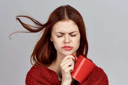 Sad red-haired girl in a red scarf and a mug on a gray background.