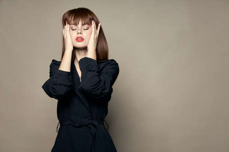 pretty brunette in a black coat with bright makeup touches her head with her hands on an isolated...