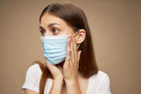 A woman in a medical mask looks to the side and touch her face with her hands on a beige background