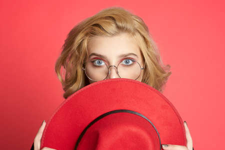 Pretty woman holds near face a red hat with glasses pink background studio