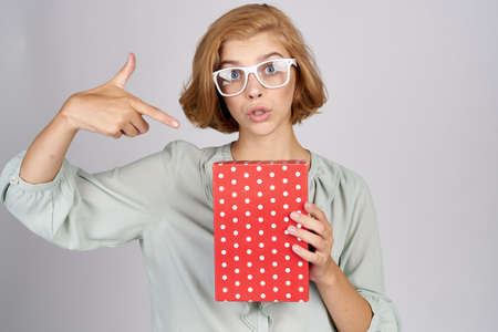 A blonde woman with a gift box gestures with her hands on a light background Copy Space