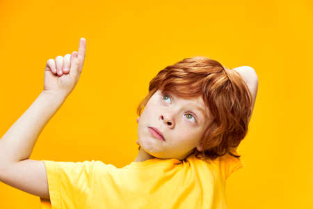 Expressive red-haired boy front view in a yellow t-shirt points up with index finger close-up, Copy...