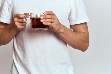 A man with a hot drink of tea in his hands in a white T-shirt on a light background cropped viewの写真素材