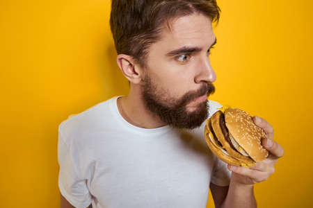 Man with hamburger in the hands of fast food diet food white t-shirt close-up yellow backgroundの写真素材