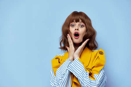 Woman in a yellow coat with an open mouth holds her hands near her face on a blue background
