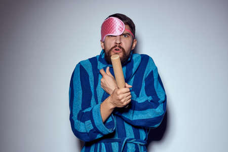 a man in a pink sleep mask holds a rolling pin in his hands and emotions energy aggression model blue robeの写真素材