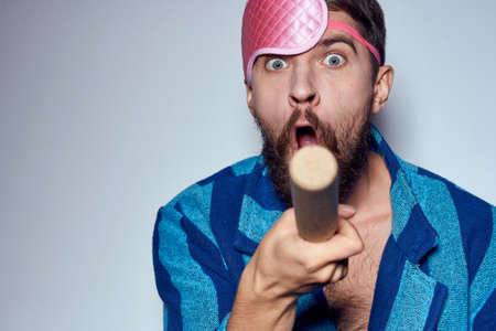 a man in a pink sleep mask holds a rolling pin in his hands and emotions energy aggression model blue robeの写真素材