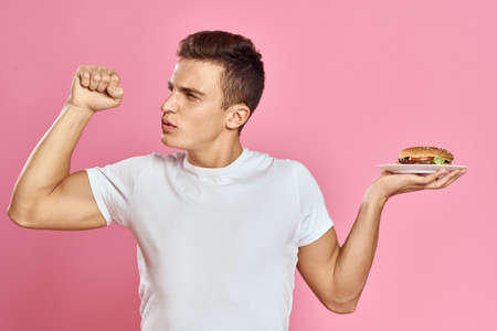 Emotional guy with hamburger on a plate and white t-shirt pink background cropped view of fast food caloriesの写真素材