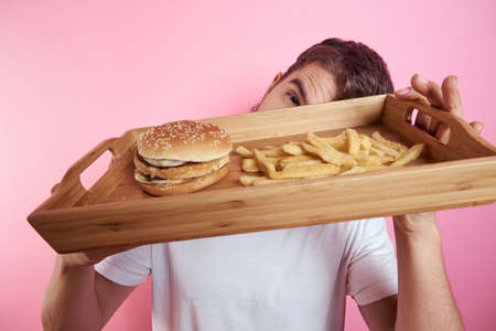 Man with a tray of food in his hand hamburger fries and fast food calorie pink background portrait close-upの写真素材