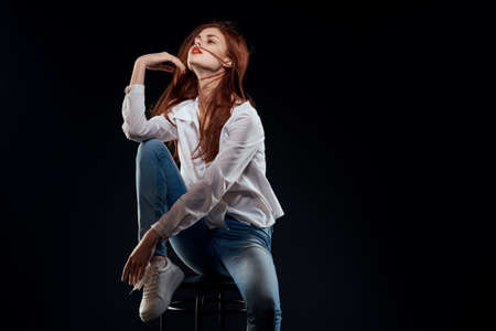 pretty woman sitting on a chair posing white shirt jeans long hair red lips dark backgroundの写真素材