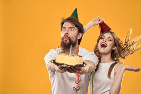 Happy man and woman in a cap celebrating a birthday on a yellow background with a cake in their handsの写真素材