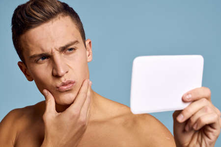 A man examines his face in a mirror on a blue background cropped viewの写真素材