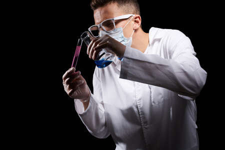 laboratory assistant in a medical mask and in a white coat with a liquid in a flask and a chemical on a black backgroundの写真素材