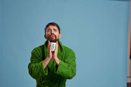 Frozen man in a warm robe with a cup of hot drink on a blue background cropped viewの写真素材