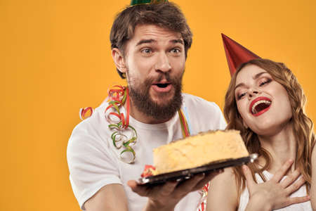 Happy man and woman in a cap celebrating a birthday on a yellow background with a cake in their...
