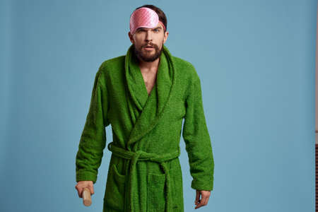 a man with a pink sleep mask holds a rolling pin in his hand and a green robe blue background emotions modelの写真素材