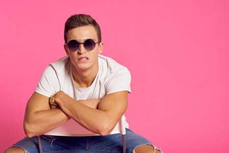 A man sits on a chair in a white t-shirt and jeans modern style pink isolated background
