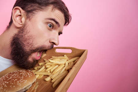 Man with a tray of food in his hand hamburger fries and fast food calorie pink background portrait close-upの写真素材