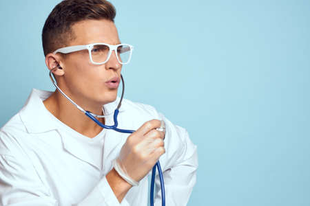 A medical worker in a dressing gown and glasses holds a stethoscope in his hands on a blue...