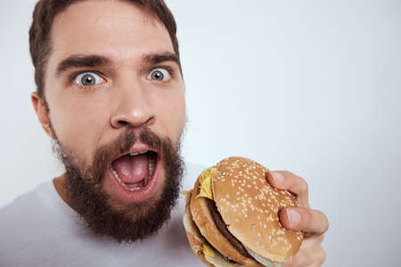 A man eating a hamburger on a light background in a white T-shirt cropped view close-up hunger fast foodの写真素材