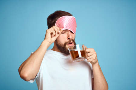 Man with pink sleep mask on his face and a cup of tea in his hands cropped view close-up blue backgroundの写真素材