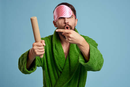 a man with a pink sleep mask holds a rolling pin in his hand and a green robe blue background emotions modelの写真素材