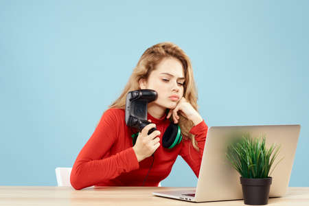 Woman sitting at a table in front of a laptop headphones controller playing online lifestyle blue...