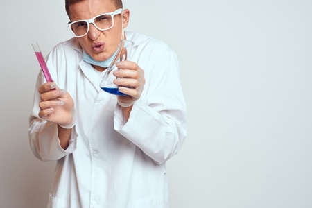A laboratory assistant in a medical gown with a protective mask holds a flask with a chemical...