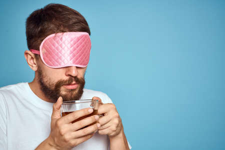 Man with pink sleep mask on his face and a cup of tea in his hands cropped view close-up blue...