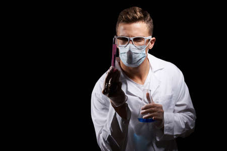 laboratory assistant in a medical mask and in a white coat with a liquid in a flask and a chemical on a black backgroundの写真素材