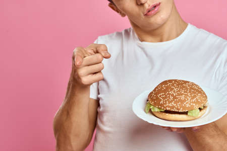 Emotional guy with hamburger on a plate and white t-shirt pink background cropped view of fast food caloriesの写真素材