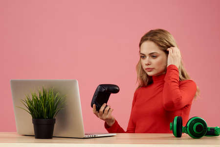 Woman sitting at a table in front of a laptop with a gamepad in hands playing technology headphones