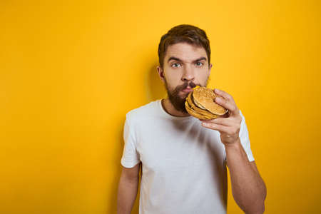 Man with hamburger in the hands of fast food diet food white t-shirt close-up yellow backgroundの写真素材
