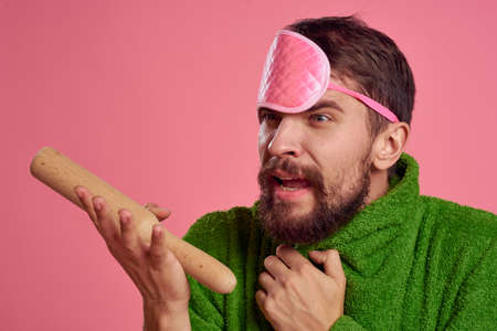 Portrait of a man in a pink sleep mask and a wooden rolling pin emotions green robe irritability modelの写真素材
