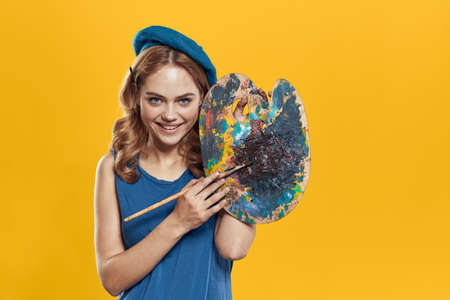 Woman with palette in blue hands takes paintbrush hobby artwork yellow background