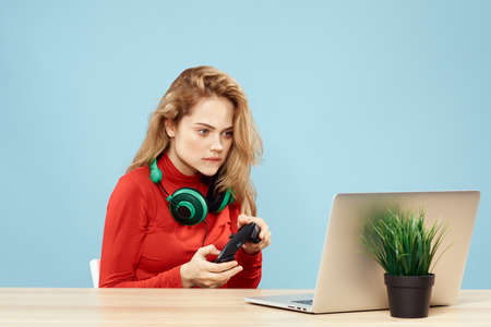 Woman sitting at a table in front of a laptop headphones controller playing online lifestyle blue backgroundの写真素材