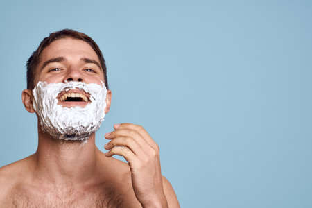 Man with razor and white foam on his face blue background nude torso bathing proceduresの写真素材