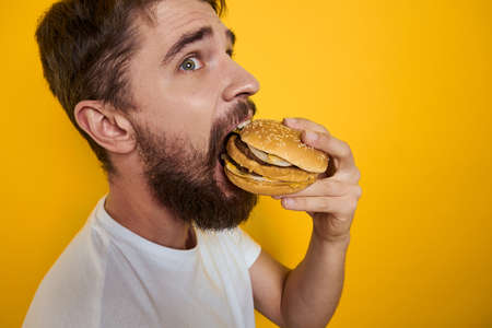 Man with hamburger in the hands of fast food diet food white t-shirt close-up yellow backgroundの写真素材