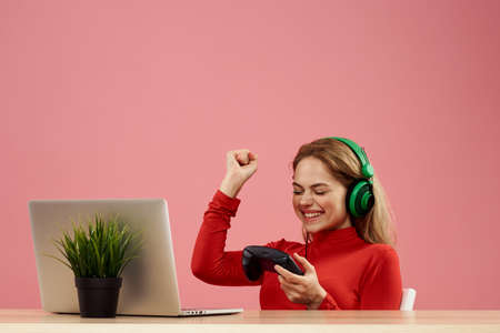 woman in headphones with gamepad in front of laptop sit at table entertainment lifestyle pink...