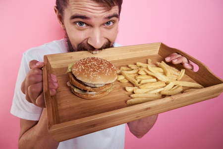 Man with a tray of food in his hand hamburger fries and fast food calorie pink background portrait close-upの写真素材