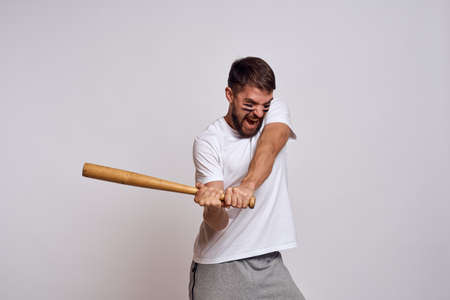 An emotional man with a baseball bat on a light background in a white T-shirt is gesturing with his...