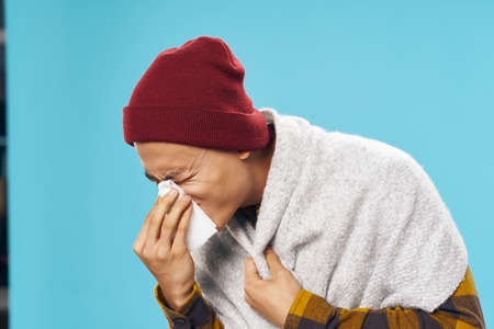 A sick man with a napkin and in warm clothes on a blue background blows his noseの写真素材