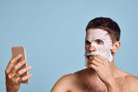 a man with a cleansing mask on his face examines himself in a mirror on a blue backgroundの写真素材