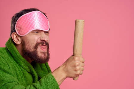 Portrait of a man in a pink sleep mask and a wooden rolling pin emotions green robe irritability...