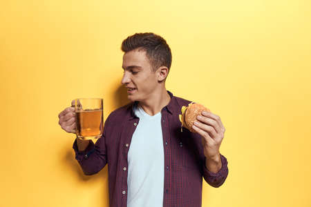Cheerful man with a mug of beer and a hamburger on his hands diet food lifestyle yellow background