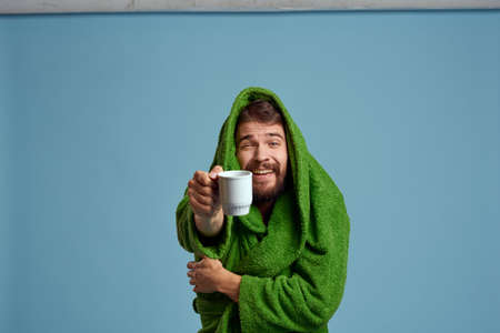 Frozen man in a warm robe with a cup of hot drink on a blue background cropped view